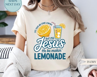 May include: Ivory-colored t-shirt featuring a graphic of a lemon, a glass of lemonade, and the text "Life gave me some lemons but my Jesus He be makin' lemonade." The shirt has short sleeves and a crew neck.