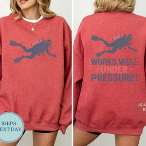 A red sweatshirt with a graphic of a scuba diver and the text "WORKS WELL UNDER PRESSURE!" printed on the back.