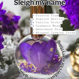 Puede incluir: Un frasco de perfume redondo y transparente con tapa dorada, que contiene una flor morada y otros elementos florales. El texto "Sleigh my name" est&aacute; en la parte superior, con notas de fragancia enumeradas a continuaci&oacute;n. Tambi&eacute;n est&aacute; presente la frase "Smells like 'my man pays for everything'".