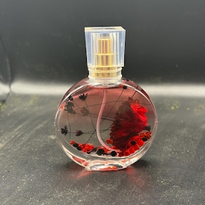 May include: A clear, round perfume bottle with a gold-coloured sprayer and a clear, square cap. The bottle contains a red liquid and decorative elements, including a large red flower and small black and red accents.