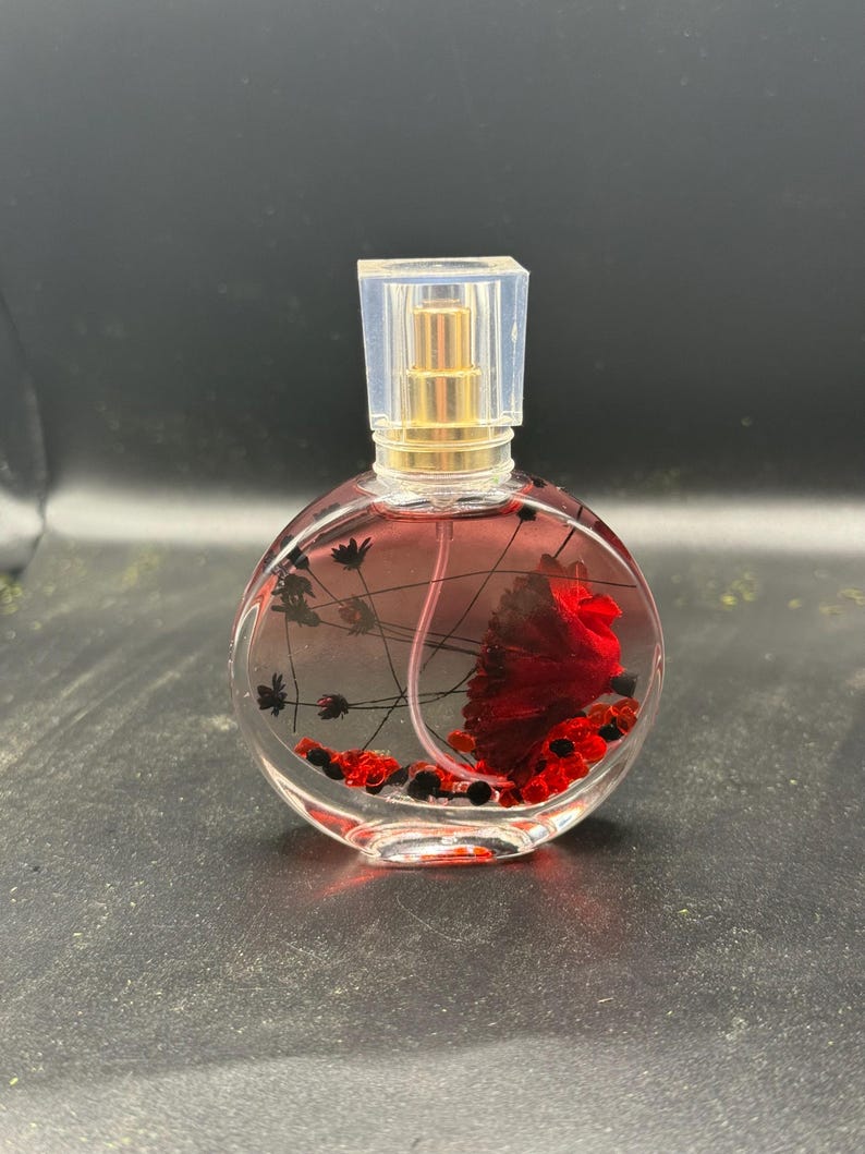 May include: A clear, round perfume bottle with a gold-coloured sprayer and a clear, square cap. The bottle contains a red liquid and decorative elements, including a large red flower and small black and red accents.