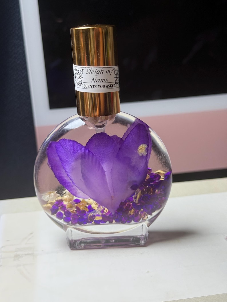 Puede incluir: Un frasco de perfume redondo y transparente con una tapa dorada y una etiqueta que dice "Sleigh my Name, Scents You Asked". El frasco contiene una gran flor morada y peque&ntilde;os elementos decorativos morados y dorados.
