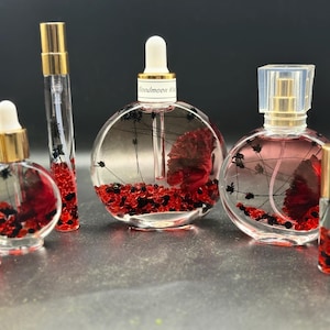 May include: A selection of clear glass perfume bottles with gold-coloured lids. The bottles are filled with red and black decorative elements, some with a floral design. The text "Bloodmoon Kiss" is visible on one bottle.