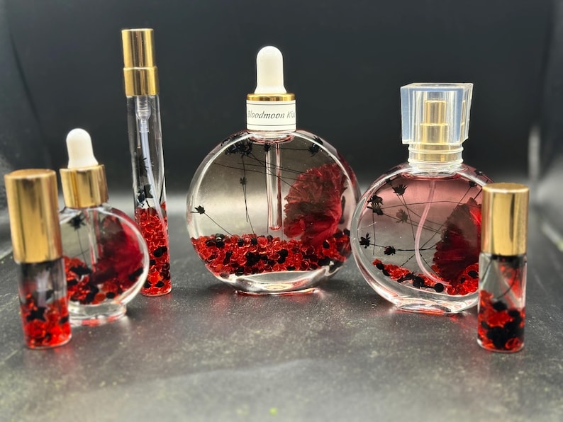 May include: A selection of clear glass perfume bottles with gold-coloured lids. The bottles are filled with red and black decorative elements, some with a floral design. The text "Bloodmoon Kiss" is visible on one bottle.