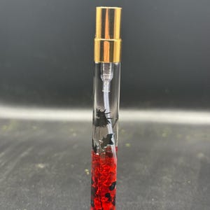 May include: A clear glass perfume vial with a gold-coloured cap. The vial contains red and black decorative elements. The perfume bottle is standing upright against a dark background.