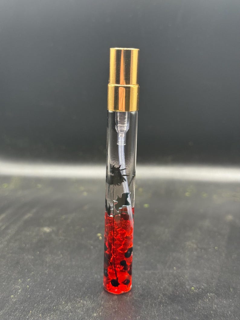 May include: A clear glass perfume vial with a gold-coloured cap. The vial contains red and black decorative elements. The perfume bottle is standing upright against a dark background.