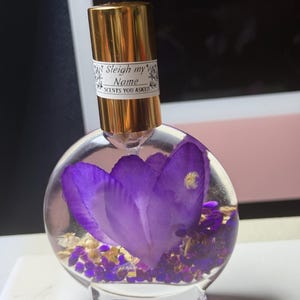 Puede incluir: Un frasco de perfume redondo y transparente con una tapa dorada y una etiqueta que dice "Sleigh my Name SCENTS YOU ASKED". El frasco contiene una gran flor morada y peque&ntilde;os elementos decorativos dorados y morados.
