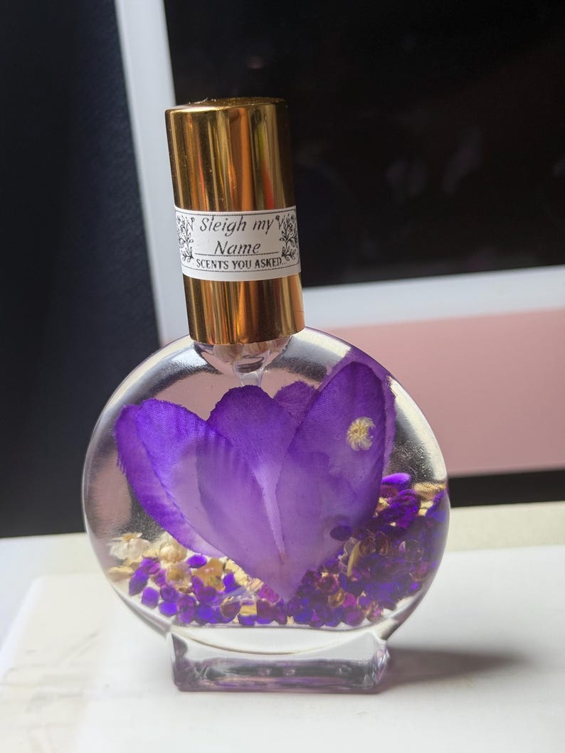 Puede incluir: Un frasco de perfume redondo y transparente con una tapa dorada y una etiqueta que dice "Sleigh my Name SCENTS YOU ASKED". El frasco contiene una gran flor morada y peque&ntilde;os elementos decorativos dorados y morados.