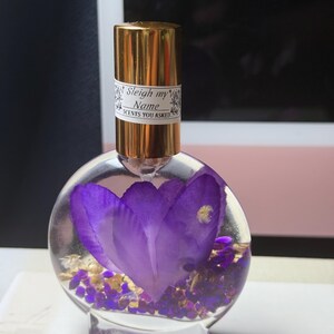 Puede incluir: Un frasco de perfume redondo y transparente con una tapa dorada y una etiqueta que dice "Sleigh my Name SCENTS YOU ASKED". El frasco contiene una gran flor morada y peque&ntilde;os detalles morados y dorados.