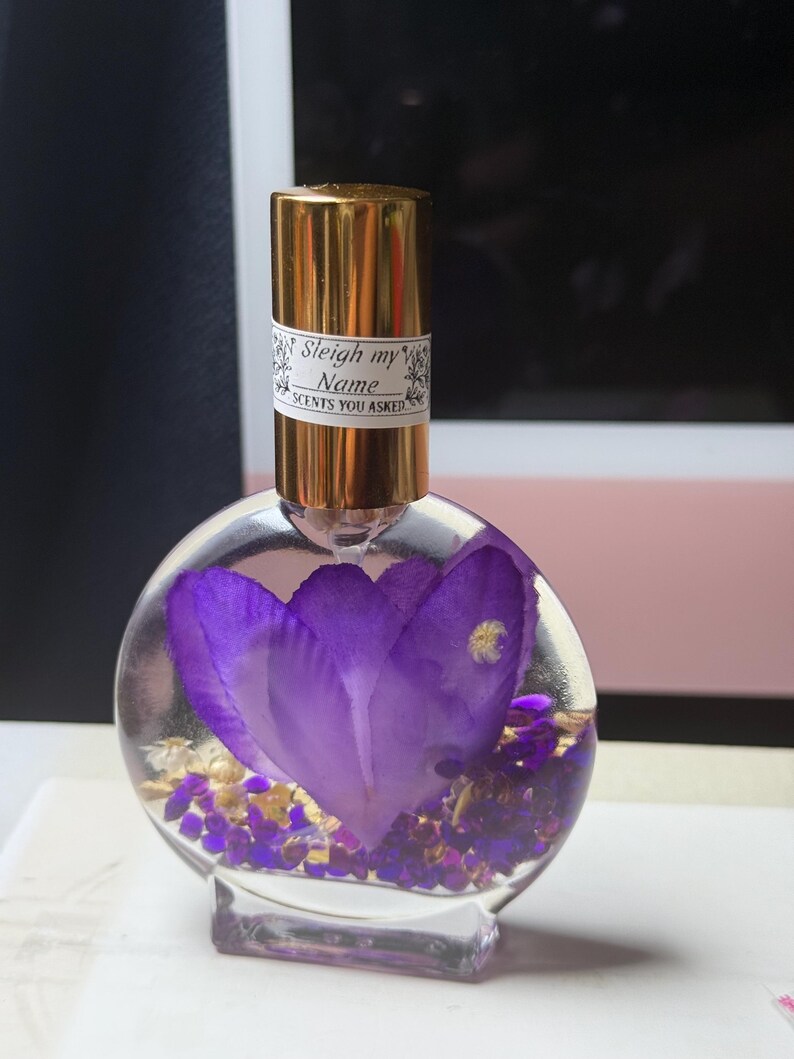 Puede incluir: Un frasco de perfume redondo y transparente con una tapa dorada y una etiqueta que dice "Sleigh my Name SCENTS YOU ASKED". El frasco contiene una gran flor morada y peque&ntilde;os detalles morados y dorados.