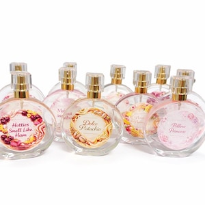 May include: A collection of clear, round perfume bottles with gold-colored caps. Each bottle has a unique label with text and decorative elements. The labels include phrases like "Hotties Smell Like Ham" and "Dolce Pistachio."