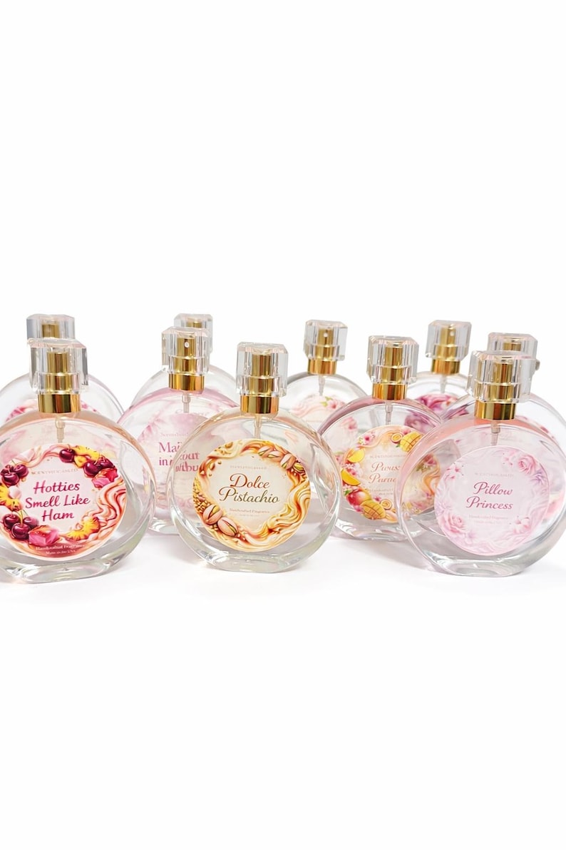May include: A collection of clear, round perfume bottles with gold-colored caps. Each bottle has a unique label with text and decorative elements. The labels include phrases like "Hotties Smell Like Ham" and "Dolce Pistachio."