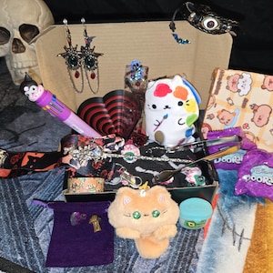 May include: A collection of various items, including earrings, a plush cat, a multi-color pen, and a small box of candy. The items are arranged in a cardboard box, with a skull visible in the background. The overall theme is a mix of cute and gothic.
