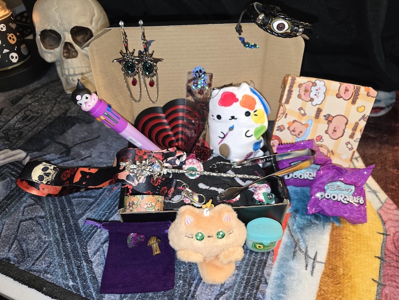 May include: A collection of various items, including earrings, a plush cat, a multi-color pen, and a small box of candy. The items are arranged in a cardboard box, with a skull visible in the background. The overall theme is a mix of cute and gothic.