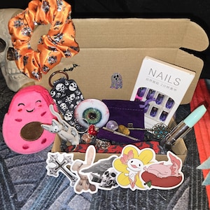 May include: A box filled with various items, including a skull, orange scrunchie, pink avocado plush, skull keychain, and a set of purple and black nails. Other items include stickers, a decorative eye, and a lipstick.