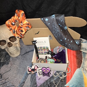 May include: A Halloween-themed gift box with a variety of items. Includes a skull, orange scrunchie with skull print, a box of "NAILS PROFESSIONAL" nail art, a black scarf with celestial design, stickers, and jewelry.