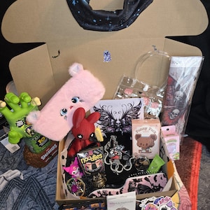 May include: An open cardboard box filled with various items, including a pink plush bunny, a red doll, and a green monster hand. Other items include stickers, a black scarf with a star pattern, and small boxes.