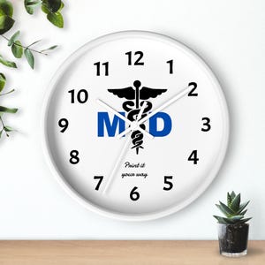 May include: White wall clock with black numerals and hands. The clock face features a black caduceus symbol above the blue letters "MD". The words "Print it your way" are printed below the letters.