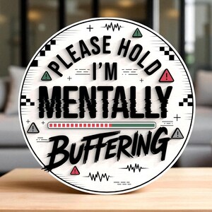 May include: A round white sign with the text "PLEASE HOLD I'M MENTALLY BUFFERING" in black, with a loading bar graphic. The sign has a black border and decorative elements, including triangles and a wavy line.