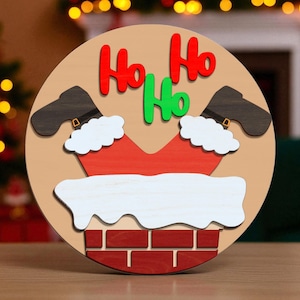 May include: A round wooden Christmas decoration featuring Santa's legs and chimney. The design includes red, green, and white elements, with the words "Ho Ho Ho" in vibrant colors. The background is a neutral tan color.