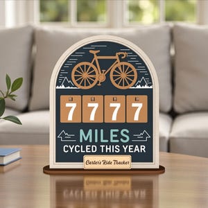 Puede incluir: Rastreador de kilómetros en bicicleta de madera con diseño de arco crema. Muestra "7777 MILES CYCLED THIS YEAR" con un gráfico de bicicleta y el texto "Carter's Ride Tracker". La paleta de colores incluye azul marino, crema y marrón.