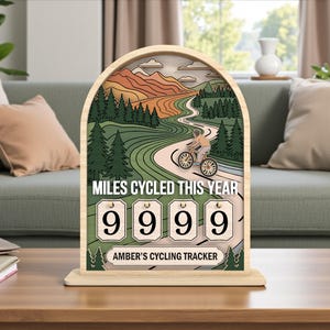 May include: Wooden cycling tracker featuring a cyclist on a winding road illustration. The text "MILES CYCLED THIS YEAR" is above the counter, displaying "9999". The tracker is personalized with "AMBER'S CYCLING TRACKER". The design is set in a wooden frame.