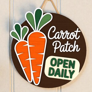 Puede incluir: Un letrero redondo de madera con fondo marrón presenta dos zanahorias naranjas con hojas verdes y las palabras "Carrot Patch" en escritura blanca. Debajo, un letrero rectangular dice "OPEN DAILY" en letras verdes.