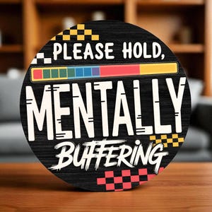 May include: Round black wooden sign with the words "PLEASE HOLD, MENTALLY BUFFERING" in white, with a colorful progress bar and checkered accents. The sign is on a wooden table.