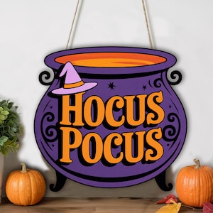 May include: A purple cauldron-shaped wooden sign with the words "Hocus Pocus" in orange. A witch hat is on the side. The sign is hanging by a string. Pumpkins and plants are in the foreground.