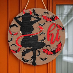 Op de afbeelding: Ronde houten Halloween-deurbord met een zwart silhouet van een duivelvrouw, rode hoorns en staart. Het bord bevat het woord "hi" in het rood, omringd door zwarte en rode vleermuizen. Er is een touw bevestigd om op te hangen.