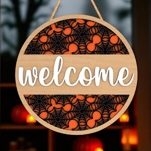 May include: Circular wooden welcome sign with a light brown finish. It features a black spiderweb design over an orange background. The word "welcome" is in white script across the centre. A rope is attached for hanging.