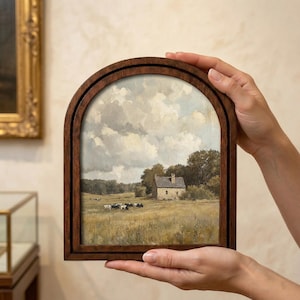 Könnte beinhalten: Ein gerahmtes Landschaftsgemälde mit einer ländlichen Szene mit einem Bauernhaus, Kühen und einem Feld unter einem bewölkten Himmel. Der Rahmen besteht aus dunklem Holz und hat eine gewölbte Oberseite. Das Gemälde wird von zwei Händen gehalten.