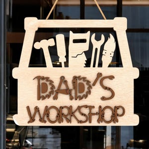 May include: Wooden sign with the words "DAD'S WORKSHOP" in brown, stylized letters. The sign features cut-out tool silhouettes, including a hammer, saw, wrench, and a figure. The sign is light brown and hangs from a natural-colored rope.