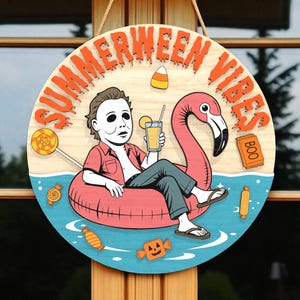 Puede incluir: Cartel de madera redondo con las palabras "SUMMERWEEN VIBES" en letras naranjas. El cartel presenta una caricatura de una persona con una máscara de Michael Myers relajándose en un flotador de flamenco rosa, sosteniendo una bebida. También se incluyen caramelos y un letrero de "BOO".
