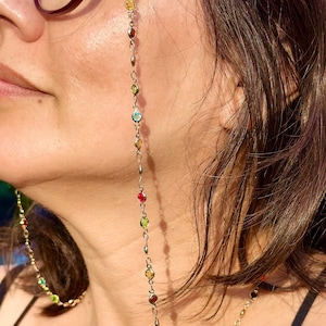 May include: A gold chain with colorful beads, worn as a glasses chain. The chain is attached to the glasses with clear plastic loops. The chain is long enough to reach the chest.