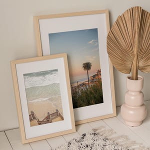 May include: Two framed beach scene prints with light wood frames lean against a wall. A decorative dried palm leaf sits in a pink vase. A white rug with black flecks and fringe is in the foreground.