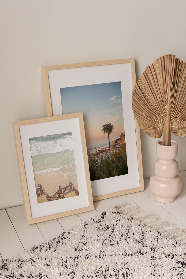 May include: Two framed beach scene prints with light wood frames lean against a wall. A decorative dried palm leaf sits in a pink vase. A white rug with black flecks and fringe is in the foreground.