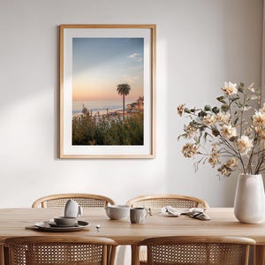 May include: A framed print of a coastal scene with a palm tree at sunset. The artwork is in a light wood frame and hangs above a wooden dining table set with dishes and a vase of flowers.