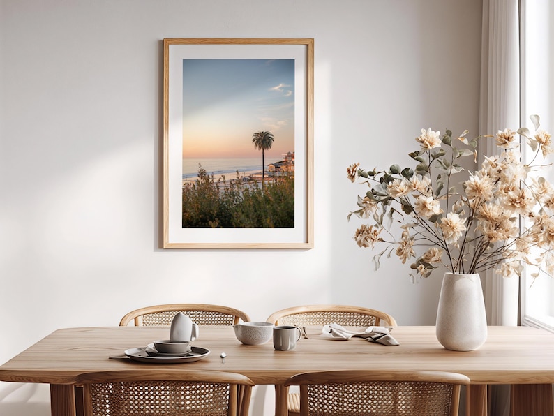 May include: A framed print of a coastal scene with a palm tree at sunset. The artwork is in a light wood frame and hangs above a wooden dining table set with dishes and a vase of flowers.