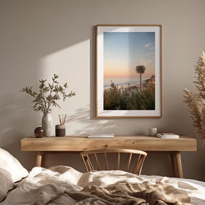 May include: A framed print of a coastal scene with a palm tree, set above a wooden desk. The print has a warm color palette of orange, blue, and green. The desk is decorated with a vase, a candle, and a small wooden bowl. A bed with neutral-colored bedding is in the foreground.