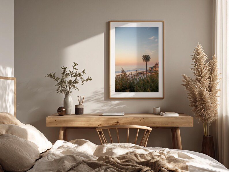 May include: A framed print of a coastal scene with a palm tree, set above a wooden desk. The print has a warm color palette of orange, blue, and green. The desk is decorated with a vase, a candle, and a small wooden bowl. A bed with neutral-colored bedding is in the foreground.