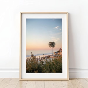 May include: Framed print of a coastal scene at sunset. The image features a palm tree, buildings, and a beach with ocean waves. The sky transitions from blue to orange and pink.
