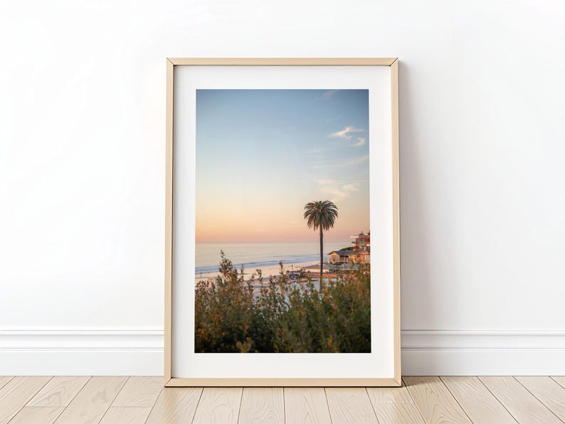 May include: Framed print of a coastal scene at sunset. The image features a palm tree, buildings, and a beach with ocean waves. The sky transitions from blue to orange and pink.