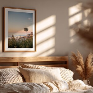 May include: A framed print of a coastal scene featuring a palm tree silhouetted against a sunset sky. The artwork is displayed above a bed with neutral-toned bedding and pillows. The room is bathed in warm sunlight.