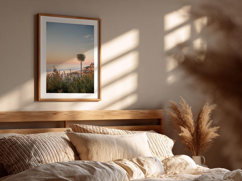 May include: A framed print of a coastal scene featuring a palm tree silhouetted against a sunset sky. The artwork is displayed above a bed with neutral-toned bedding and pillows. The room is bathed in warm sunlight.