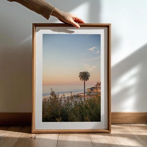 May include: A framed print of a coastal landscape featuring a palm tree silhouetted against a sunset sky. The image captures a beach scene with buildings and vegetation, framed in a natural wood. The print is held by a person's hand.