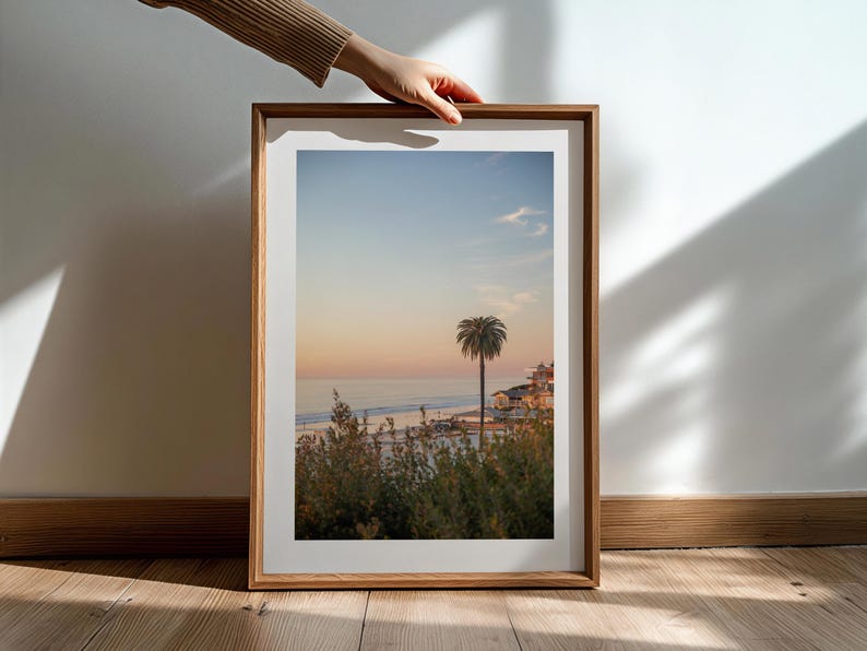 May include: A framed print of a coastal landscape featuring a palm tree silhouetted against a sunset sky. The image captures a beach scene with buildings and vegetation, framed in a natural wood. The print is held by a person's hand.
