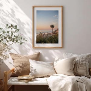 May include: A framed print of a coastal scene with a palm tree at sunset. The artwork is displayed above a bench with beige and white pillows and a blanket. A potted plant is to the left.
