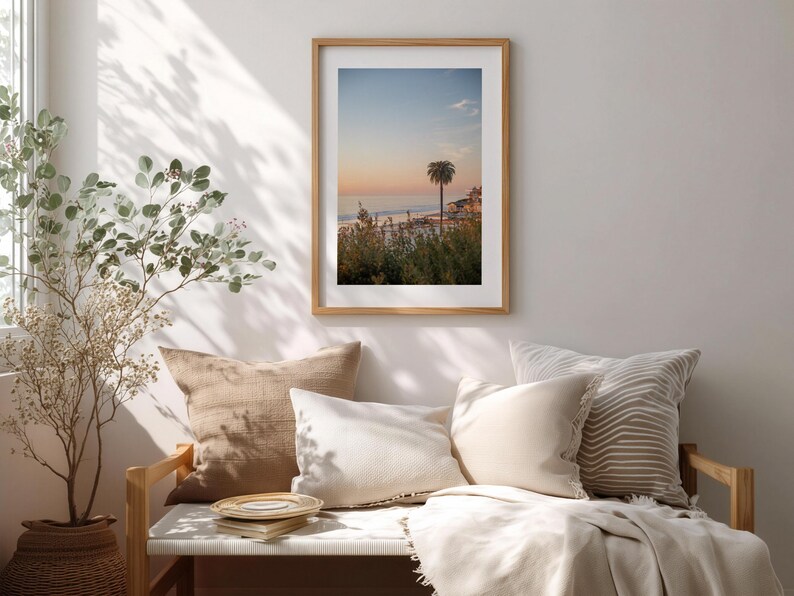 May include: A framed print of a coastal scene with a palm tree at sunset. The artwork is displayed above a bench with beige and white pillows and a blanket. A potted plant is to the left.