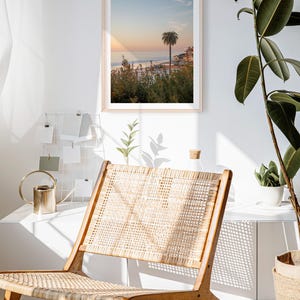 May include: A framed print of a coastal scene with a palm tree at sunset hangs on a white wall. A wooden chair with a woven seat and back sits in front of the wall. A gold watering can and a plant are on a white shelf.